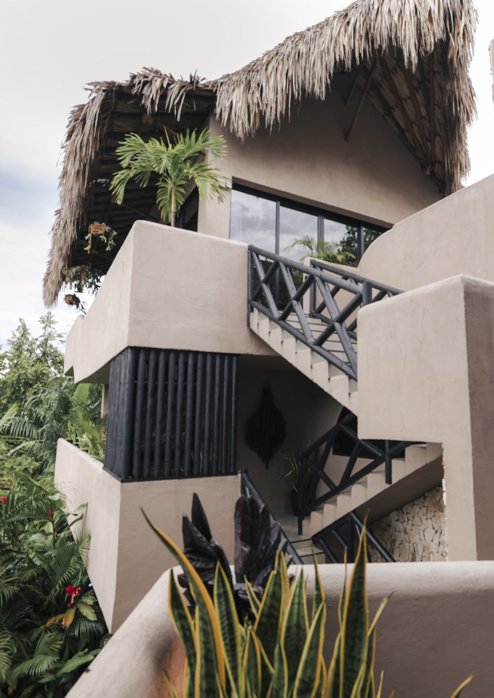 Two-story tropical-style building with a thatched roof, beige stucco walls, and a metal staircase among lush plants.