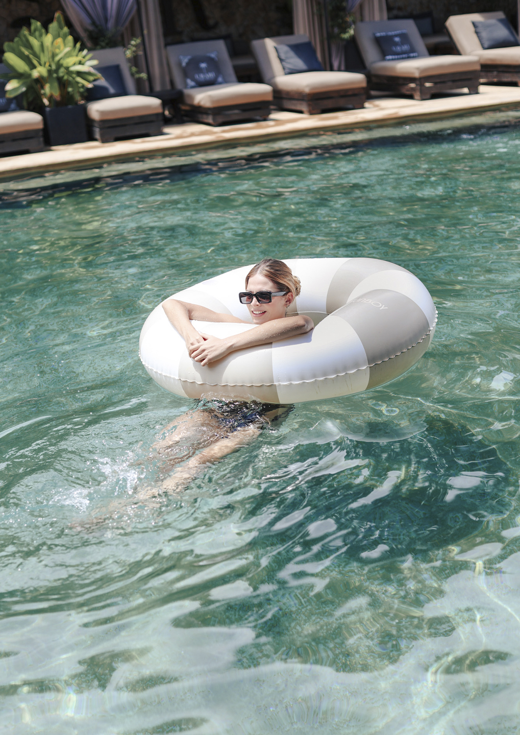 Woman wearing sunglasses relaxing in a striped inflatable pool ring, floating in a clear turquoise pool with resort lounge chairs in the background.