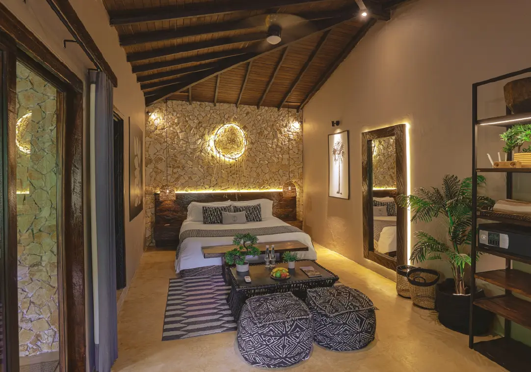 A cozy hotel bedroom with a stone feature wall, circular wall light, and a low wooden headboard; bed with white linens and patterned throw pillows sits opposite a glass coffee table and woven ottomans on a geometric rug.