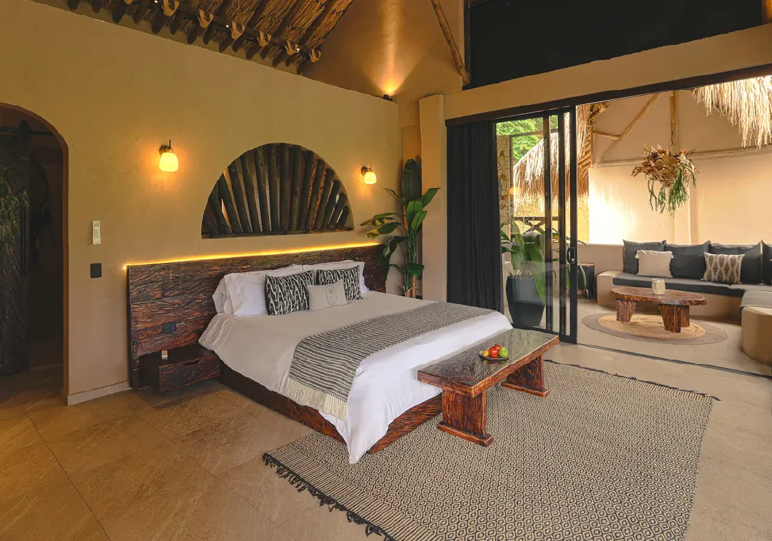 Luxurious bedroom with a king bed, white linens, dark wood headboard, and a wooden bench at the foot, softly lit by wall lamps; open doorway to a lounge area.