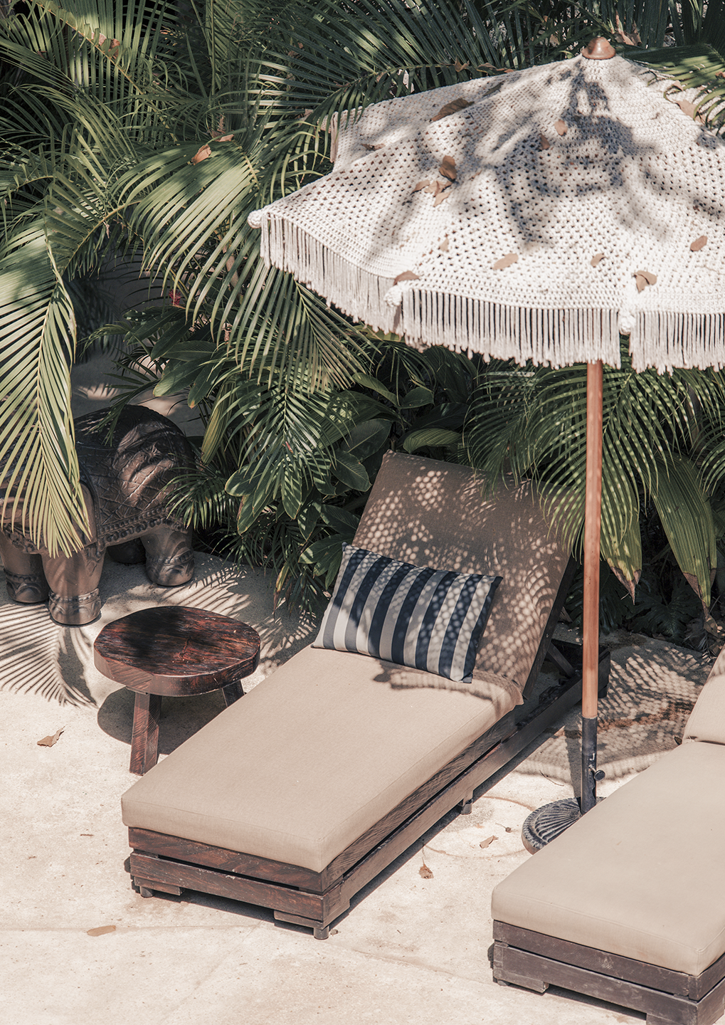 Outdoor tropical seating area with a beige cushioned lounge chair, striped pillow, round wooden table, and a fringed parasol among palm leaves.