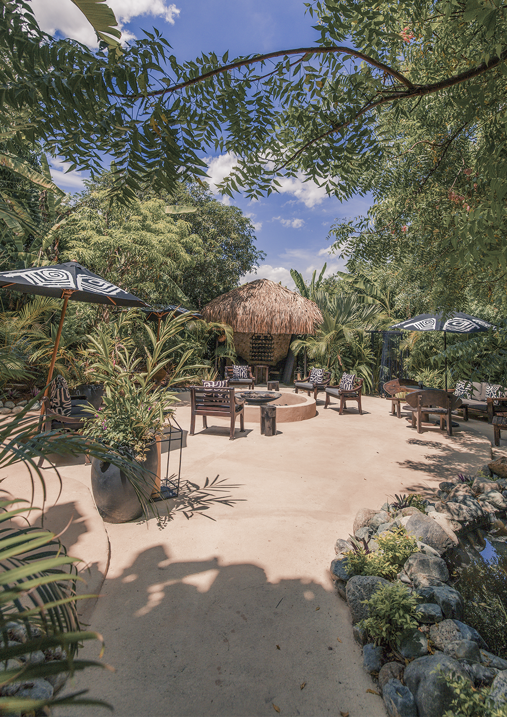 Outdoor tropical lounge with a thatched hut, circular fire pit, and wicker chairs under patterned umbrellas.